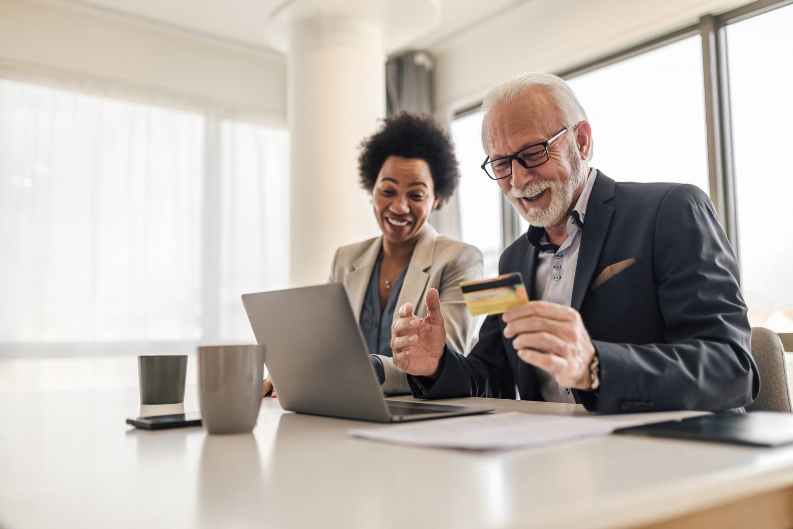 Happy male and female colleagues paying online through credit card. Senior and young professionals making transactions on laptop. They are working at corporate office.