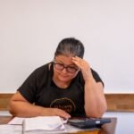 Adult woman looks stressed while working on finances with calculator and notes at a desk.