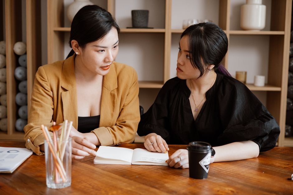 Empreendedorismo na prática: como transformar uma ideia em um negócio consistente 2 Two women engaged in a business meeting at an office desk with notebooks and coffee.