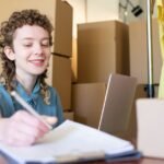 Smiling young woman managing a small business with laptop and packages around her.