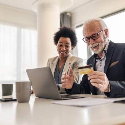 Happy male and female colleagues paying online through credit card. Senior and young professionals making transactions on laptop. They are working at corporate office.